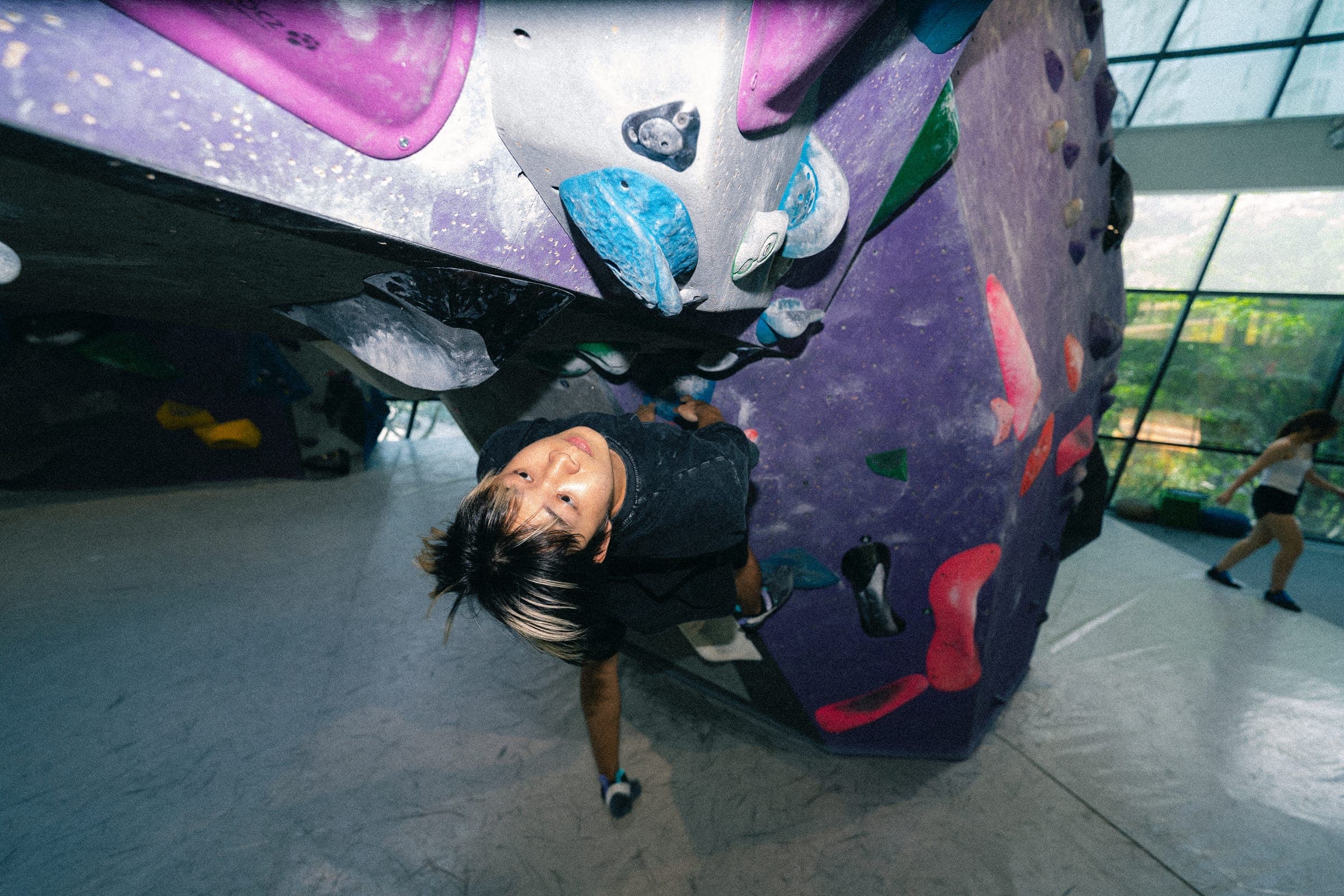 A climber inverted under a purple roof, looking back toward the camera.